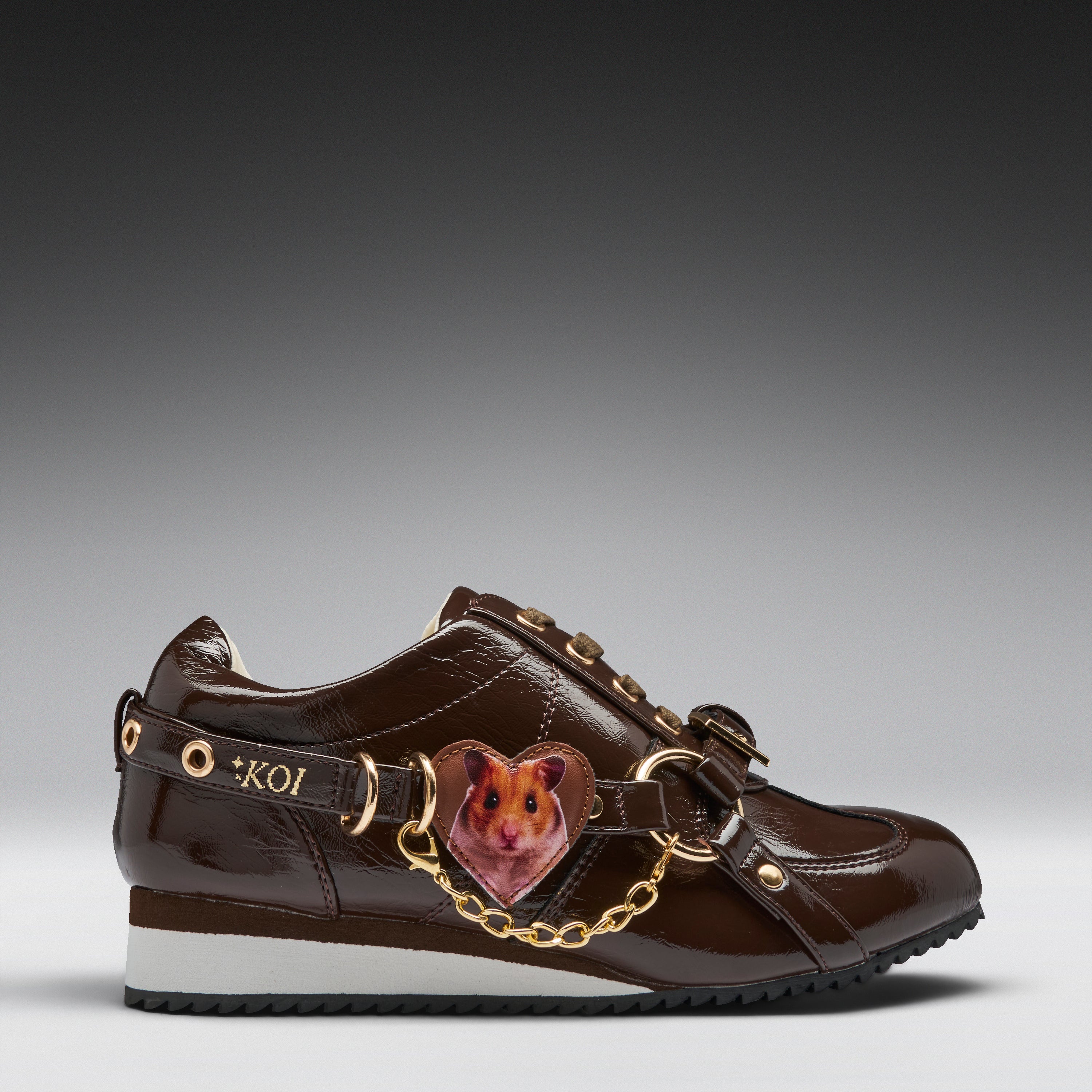 A brown patent leather sneaker featuring a heart-shaped hamster charm attached by a gold chain and strap, with 'KOI' lettering on the side.