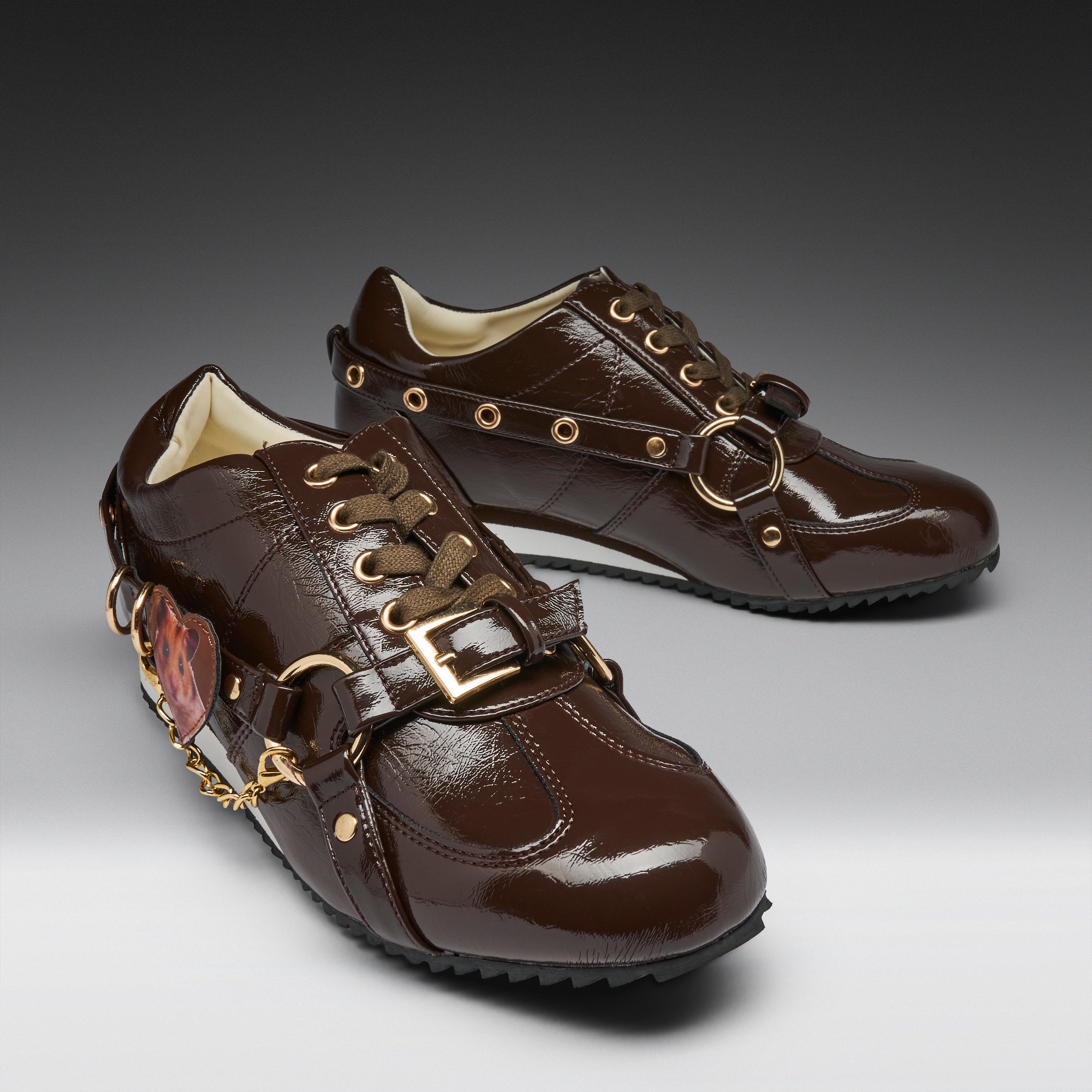 A pair of dark brown patent leather lace-up shoes with decorative gold-tone hardware, including buckles, rings, eyelets, and a dangling chain with a heart-shaped charm, shown on a gray gradient background.