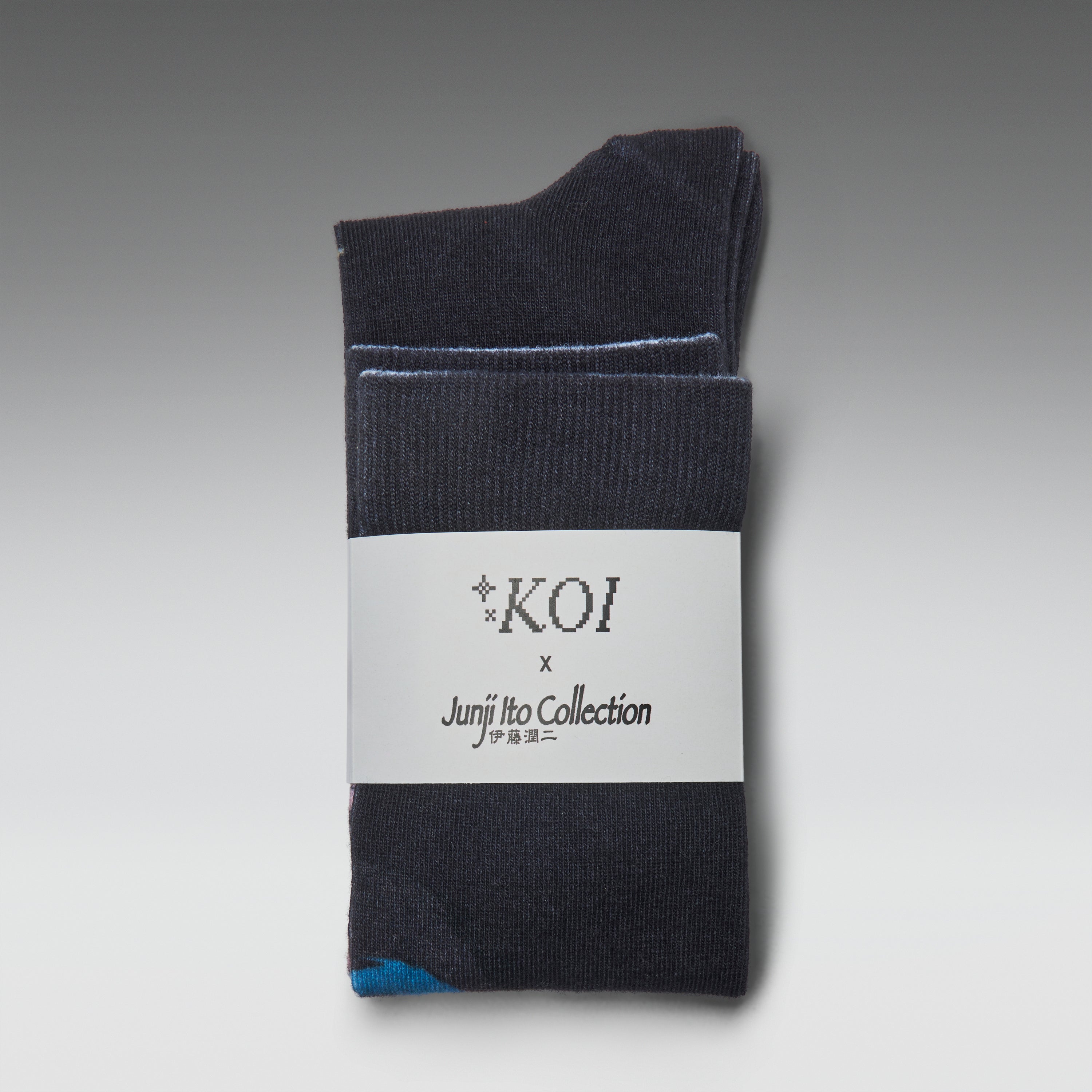 A pair of dark charcoal gray crew socks folded together with a white paper label that reads 'KOI x Junji Ito Collection'. A small light blue accent is visible on the toe area.