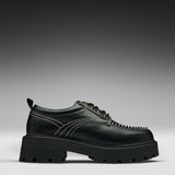 A black leather lace-up boot with a chunky lug sole, contrast white stitching, and gold-tone eyelets, shown in a side profile on a gray gradient background.