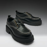 A pair of black leather lace-up shoes with chunky lug soles, contrast white stitching, and gold-tone eyelets, displayed on a dark-to-light gray gradient studio background.