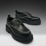 A pair of black leather lace-up shoes with chunky lug soles, contrast white stitching, and gold-tone eyelets, displayed on a dark-to-light gray gradient studio background.