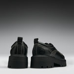 A pair of chunky black platform ankle boots with dark iridescent panels, side buckle straps, and a small circular logo on the heel, shown from the back and side against a dark gray gradient background.