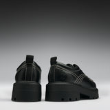 A pair of chunky black platform ankle boots with dark iridescent panels, side buckle straps, and a small circular logo on the heel, shown from the back and side against a dark gray gradient background.