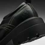 Close-up of a black leather shoe heel showing a circular embossed logo, white contrast stitching, and a chunky black sole on a dark gray gradient background.
