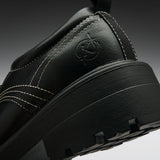 Close-up of a black leather shoe heel showing a circular embossed logo, white contrast stitching, and a chunky black sole on a dark gray gradient background.