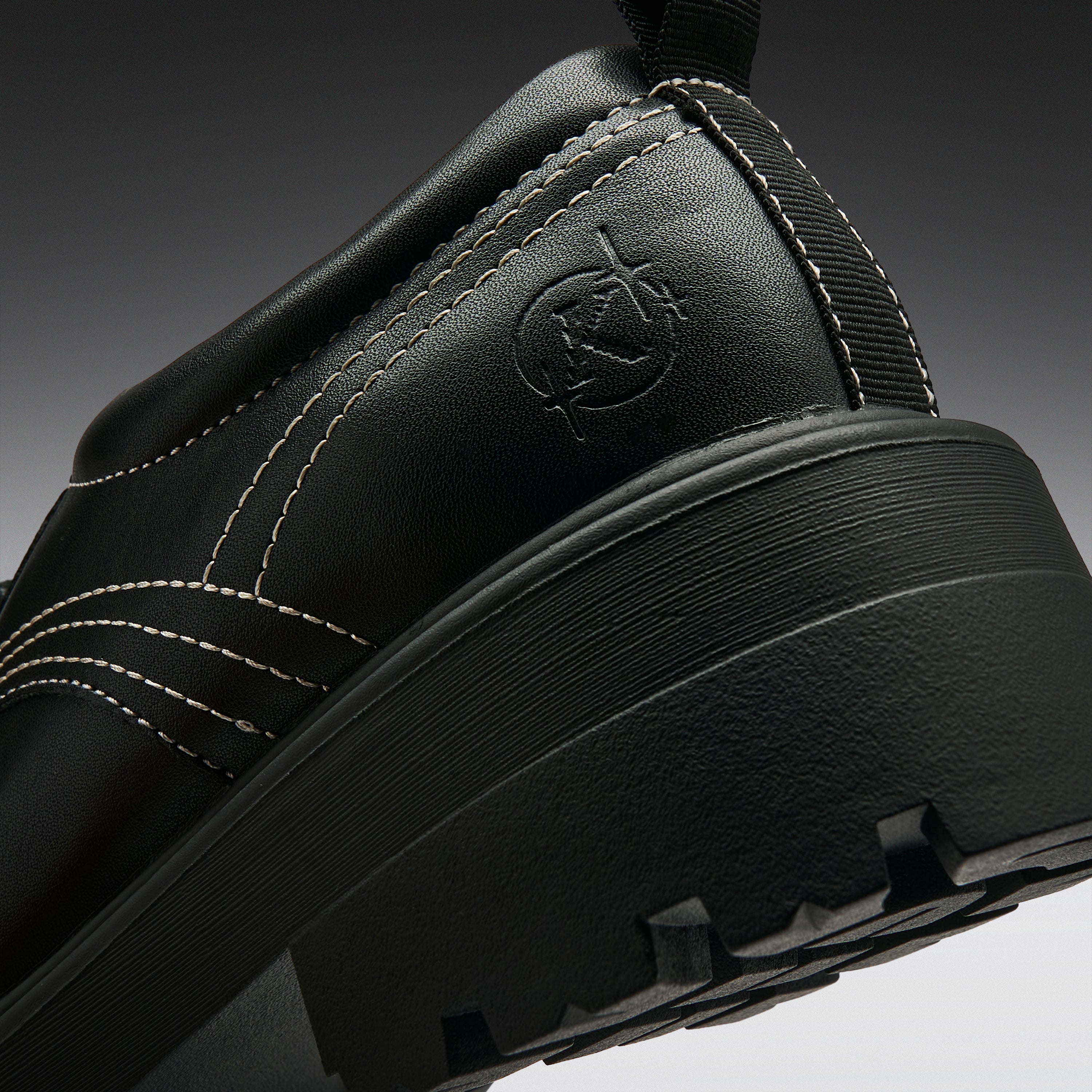 Close-up of a black leather shoe heel showing a circular embossed logo, white contrast stitching, and a chunky black sole on a dark gray gradient background.