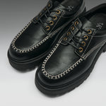 A pair of black leather lace-up shoes with distinctive white contrast stitching along the toe, gold-tone metal eyelets, and thick black soles, shown on a light gray gradient background.