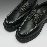A pair of black leather lace-up shoes with distinctive white contrast stitching along the toe, gold-tone metal eyelets, and thick black soles, shown on a light gray gradient background.