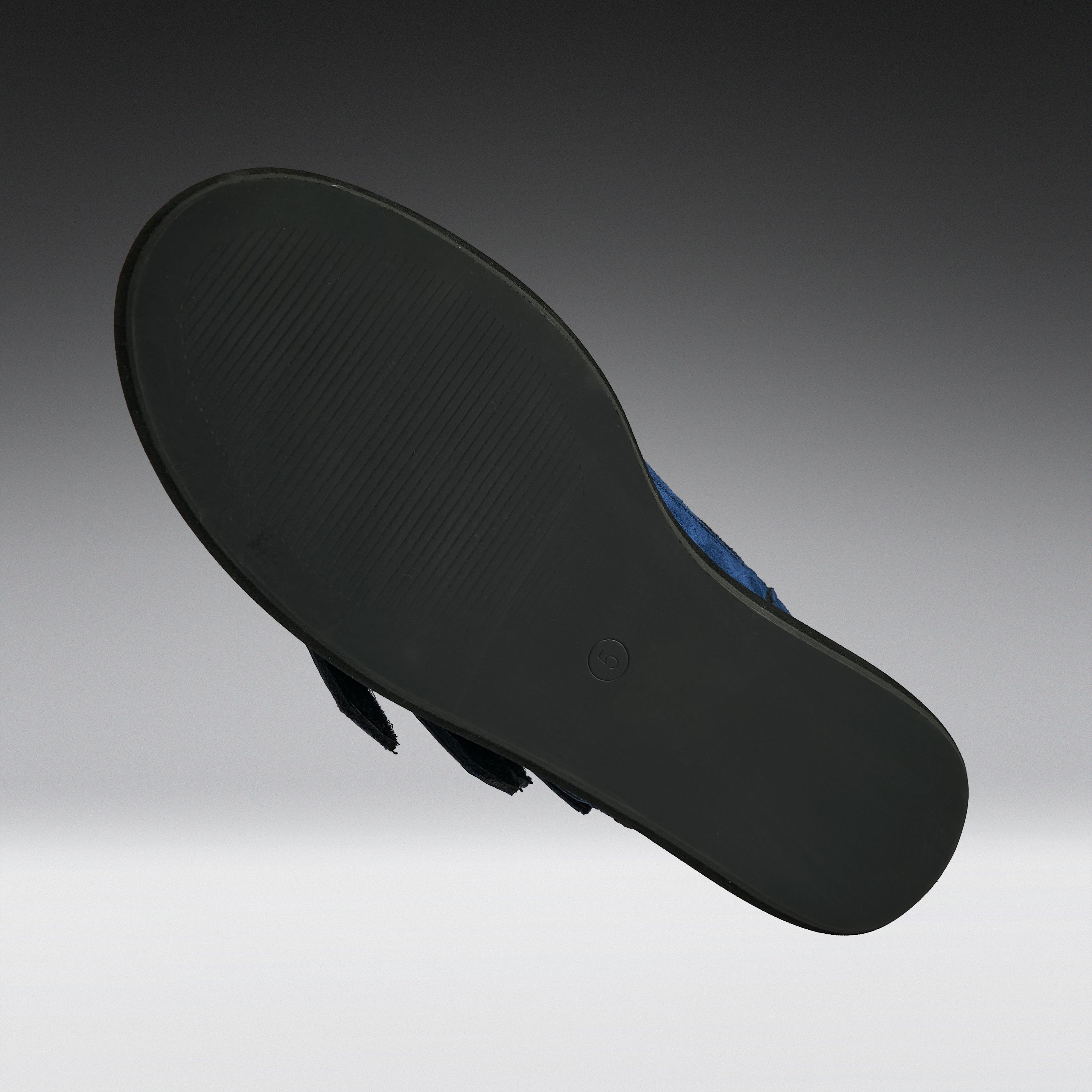 The black outsole of a flat sandal, shown floating against a dark gray to light gray gradient background. A small portion of the blue upper is visible on the right side.