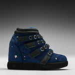 A high-top sneaker in dark blue suede and black faux leather, featuring multiple adjustable straps with silver-tone metal rings, star embellishments, and a thick black sole, photographed on a gray gradient studio background.