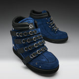 A pair of blue suede and black leather high-top boots with multiple black straps and silver metal O-rings, shown on a gray gradient background.
