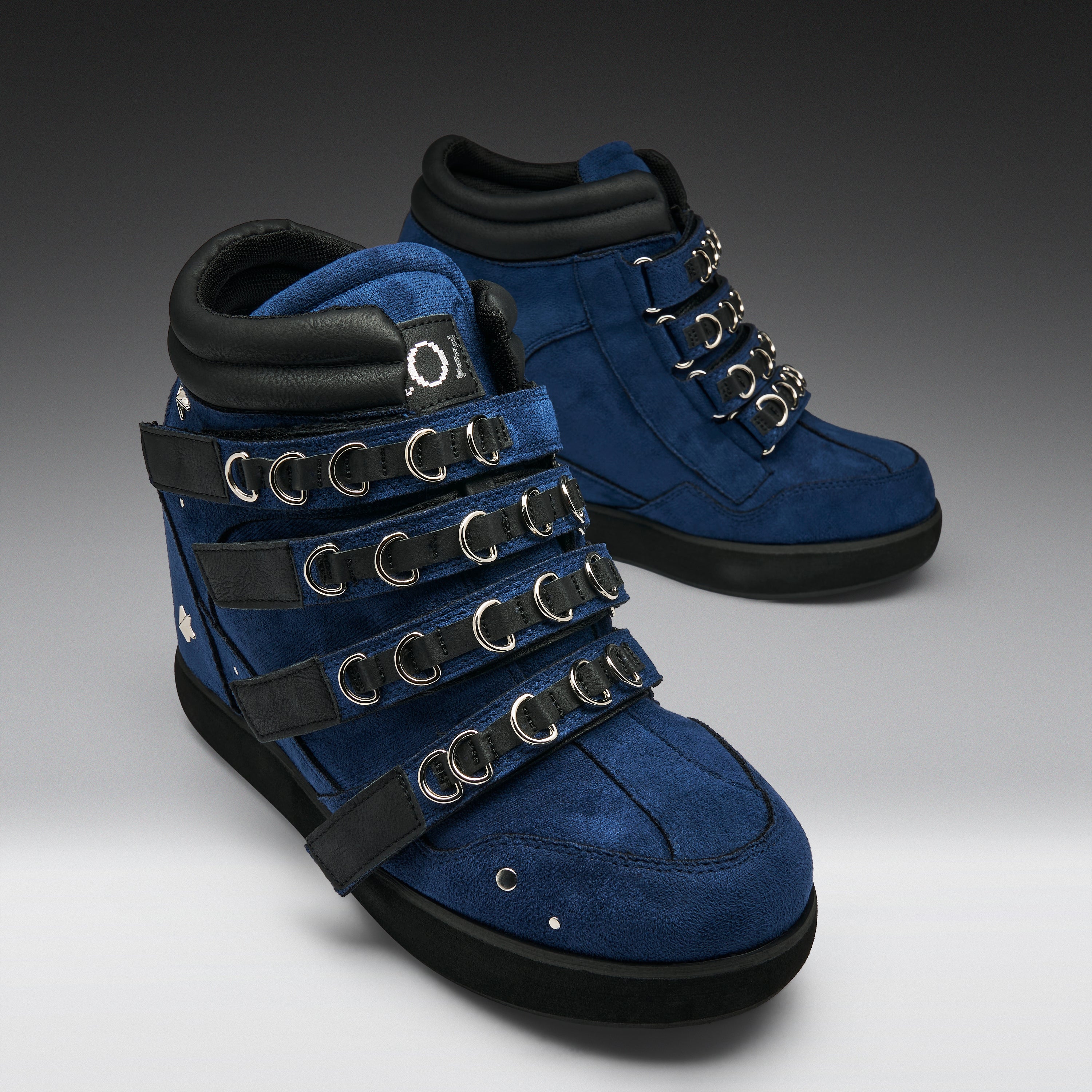 A pair of blue suede and black leather high-top boots with multiple black straps and silver metal O-rings, shown on a gray gradient background.