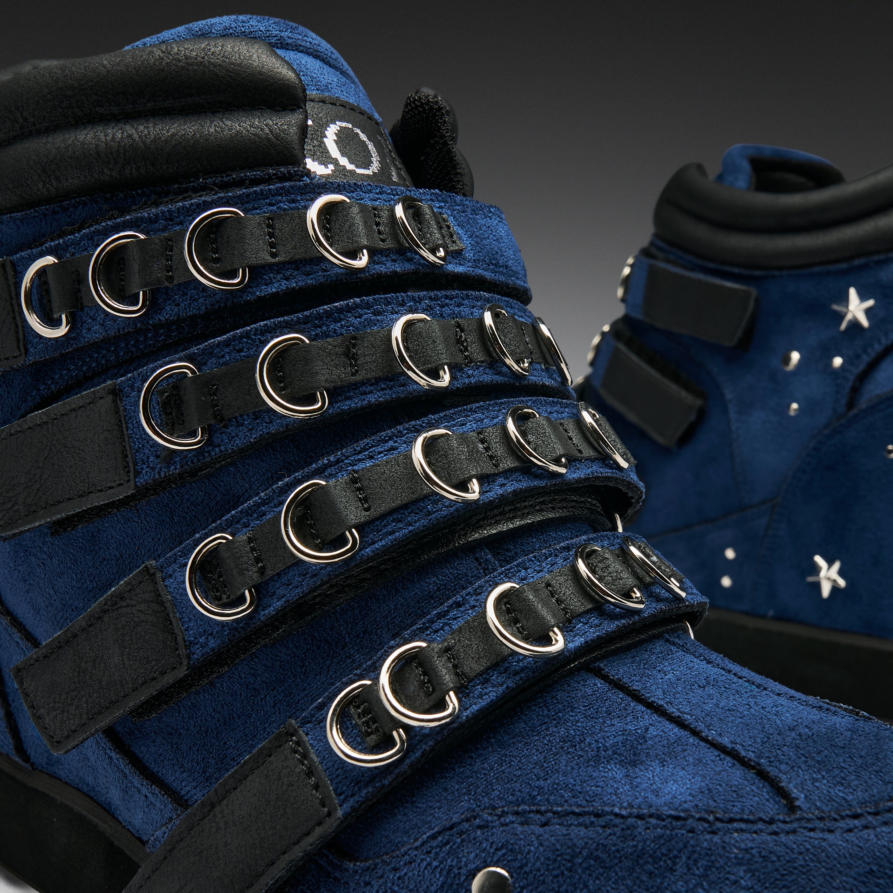 A close-up of a blue suede high-top sneaker with multiple black straps, silver metal D-rings, and small white star embellishments on the side.