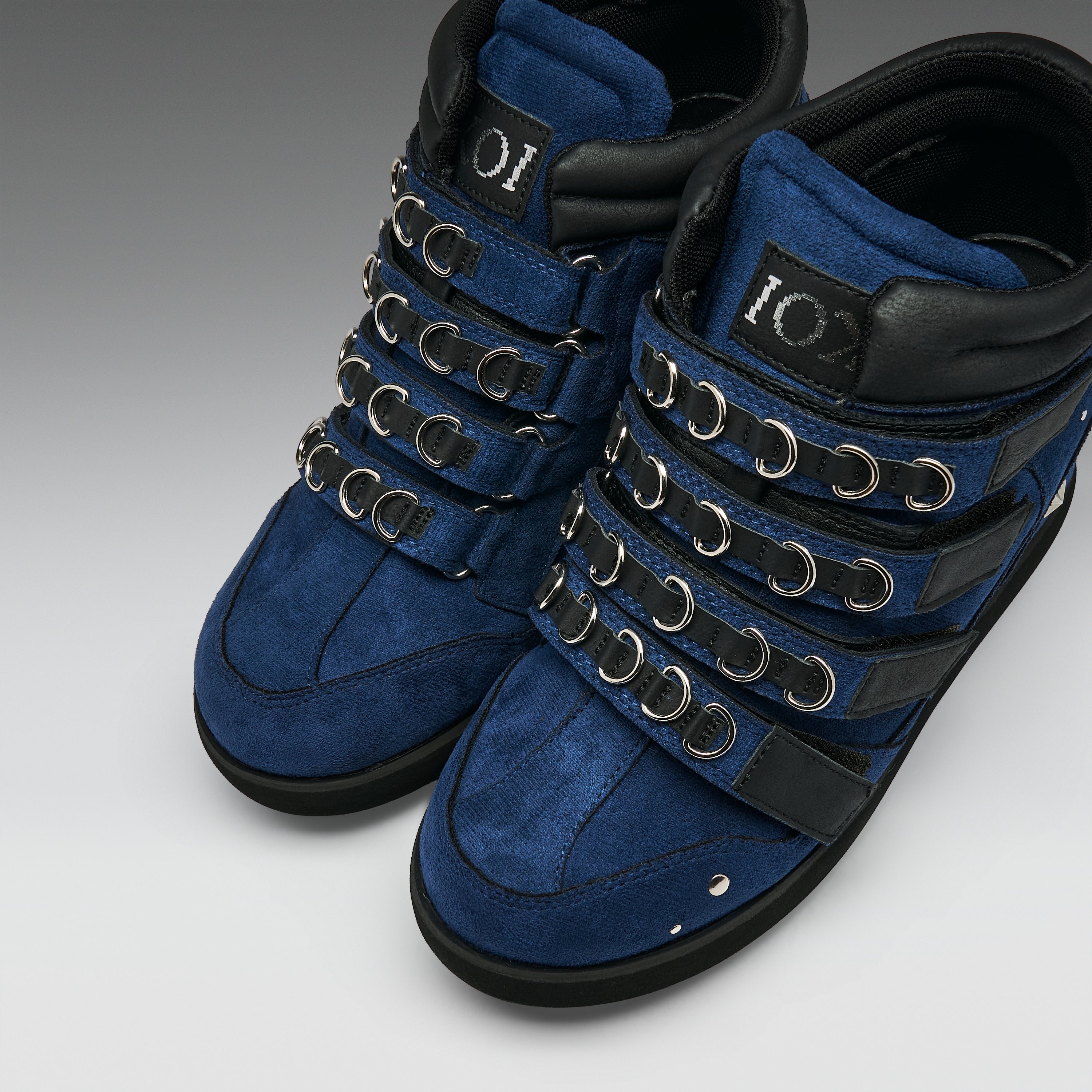 A pair of high-top blue suede boots with black leather trim, multiple rows of silver metal eyelets, and a black sole, shown on a neutral gray background.
