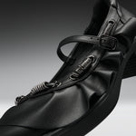 A close-up studio shot of a black leather Mary Jane-style shoe with multiple spring-loaded metal loops and a buckle strap, set against a dark gray gradient background.