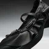 A close-up studio shot of a black leather Mary Jane-style shoe with multiple spring-loaded metal loops and a buckle strap, set against a dark gray gradient background.
