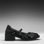 A single black leather Mary Jane-style shoe with a chunky block heel, decorative ruffles, and a strap with a metallic ring detail, shown in profile against a dark gray gradient background.