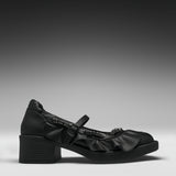 A single black leather Mary Jane-style shoe with a chunky block heel, decorative ruffles, and a strap with a metallic ring detail, shown in profile against a dark gray gradient background.