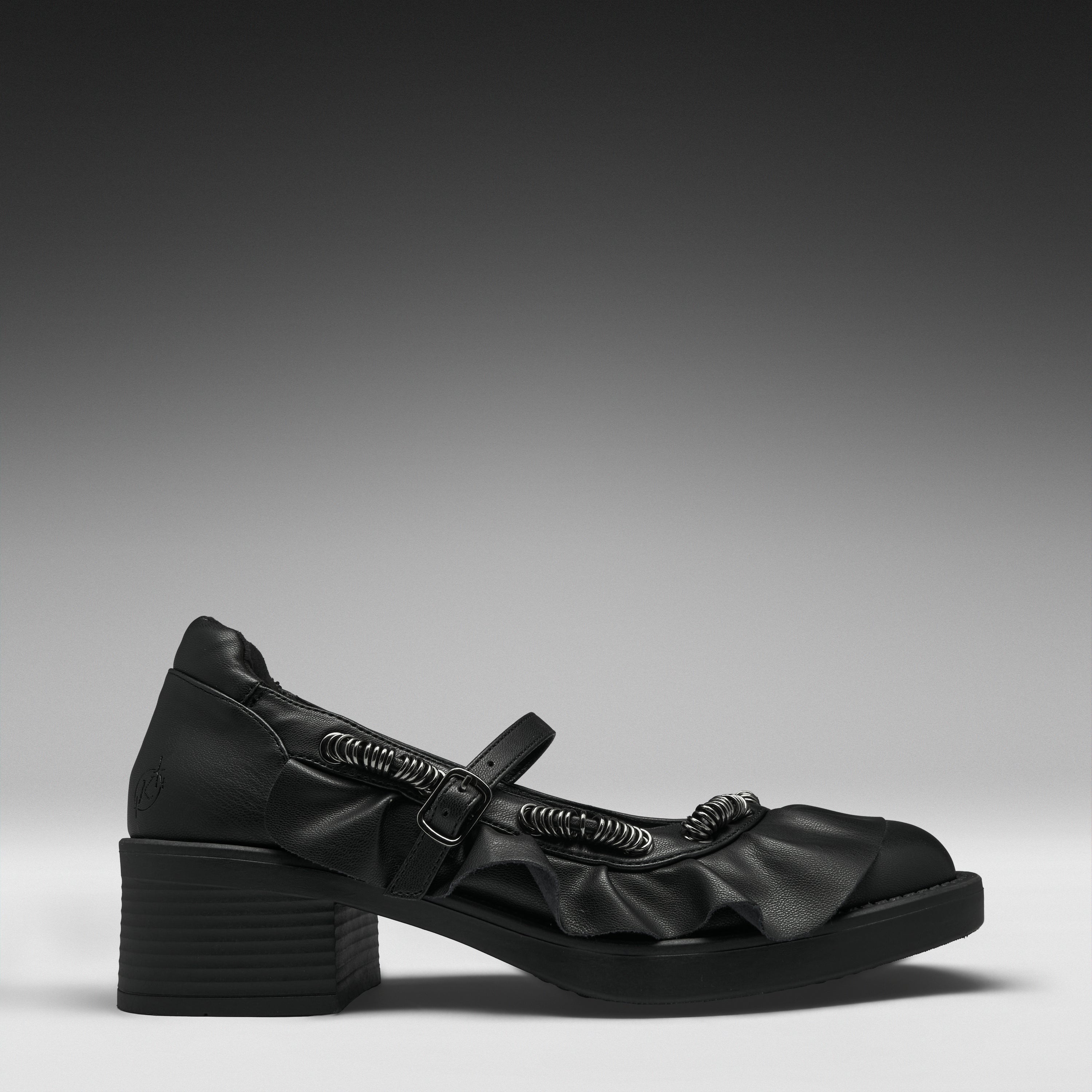 A single black leather Mary Jane-style shoe with a chunky block heel, decorative ruffles, and a strap with a metallic ring detail, shown in profile against a dark gray gradient background.