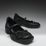 A pair of black Mary Jane-style shoes with chunky block heels, ruffled straps, and silver-tone decorative hardware, photographed on a dark-to-light gray studio background.