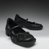 A pair of black Mary Jane-style shoes with chunky block heels, ruffled straps, and silver-tone decorative hardware, photographed on a dark-to-light gray studio background.