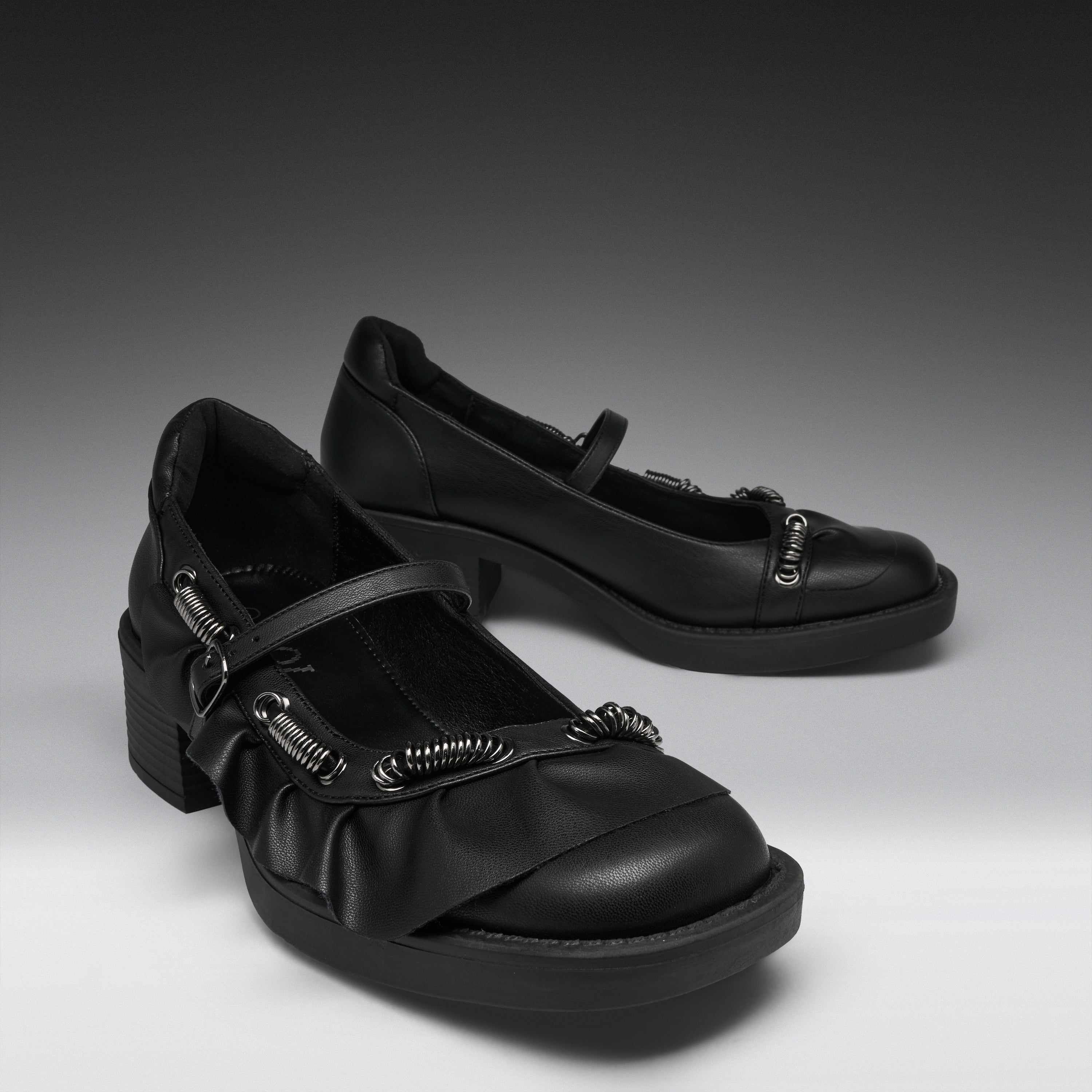 A pair of black Mary Jane-style shoes with chunky block heels, ruffled straps, and silver-tone decorative hardware, photographed on a dark-to-light gray studio background.