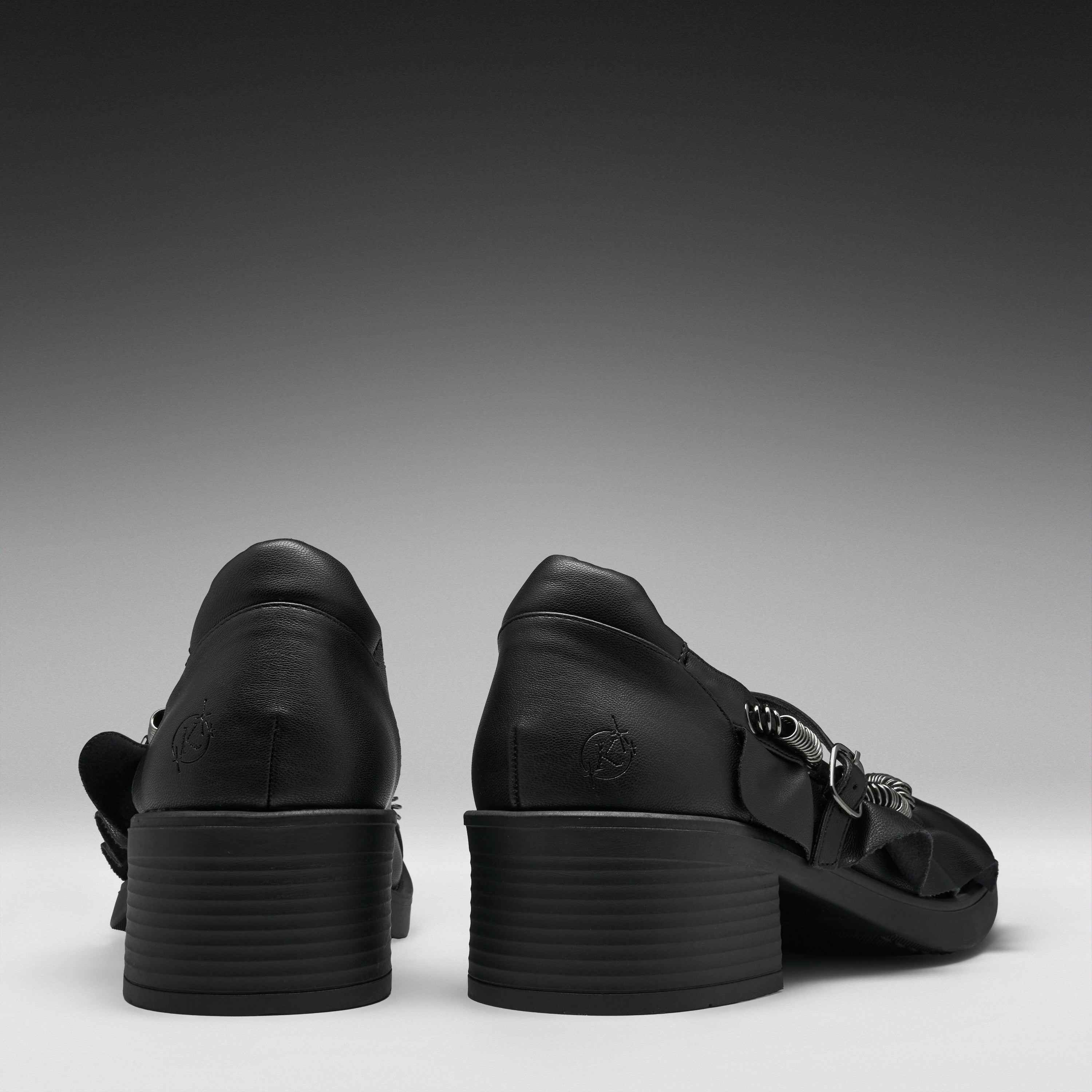 A pair of black leather high-heeled sandals shown from the back, featuring chunky stacked block heels and decorative side straps, set against a dark gray gradient background.