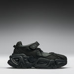 A black chunky platform sneaker with a hook-and-loop strap closure, shown from a side profile against a dark gray gradient studio background.