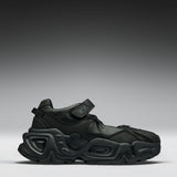 A black chunky platform sneaker with a hook-and-loop strap closure, shown from a side profile against a dark gray gradient studio background.