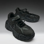 A pair of chunky black sneakers with a textured leather upper, thick sculpted black soles, and hook-and-loop strap closures, displayed on a dark-to-light gray gradient studio background.