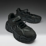 A pair of chunky black sneakers with a textured leather upper, thick sculpted black soles, and hook-and-loop strap closures, displayed on a dark-to-light gray gradient studio background.
