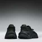 A pair of black chunky platform sneakers shown from the back, positioned side-by-side against a dark gray to light gray gradient studio background.