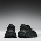 A pair of black chunky platform sneakers shown from the back, positioned side-by-side against a dark gray to light gray gradient studio background.