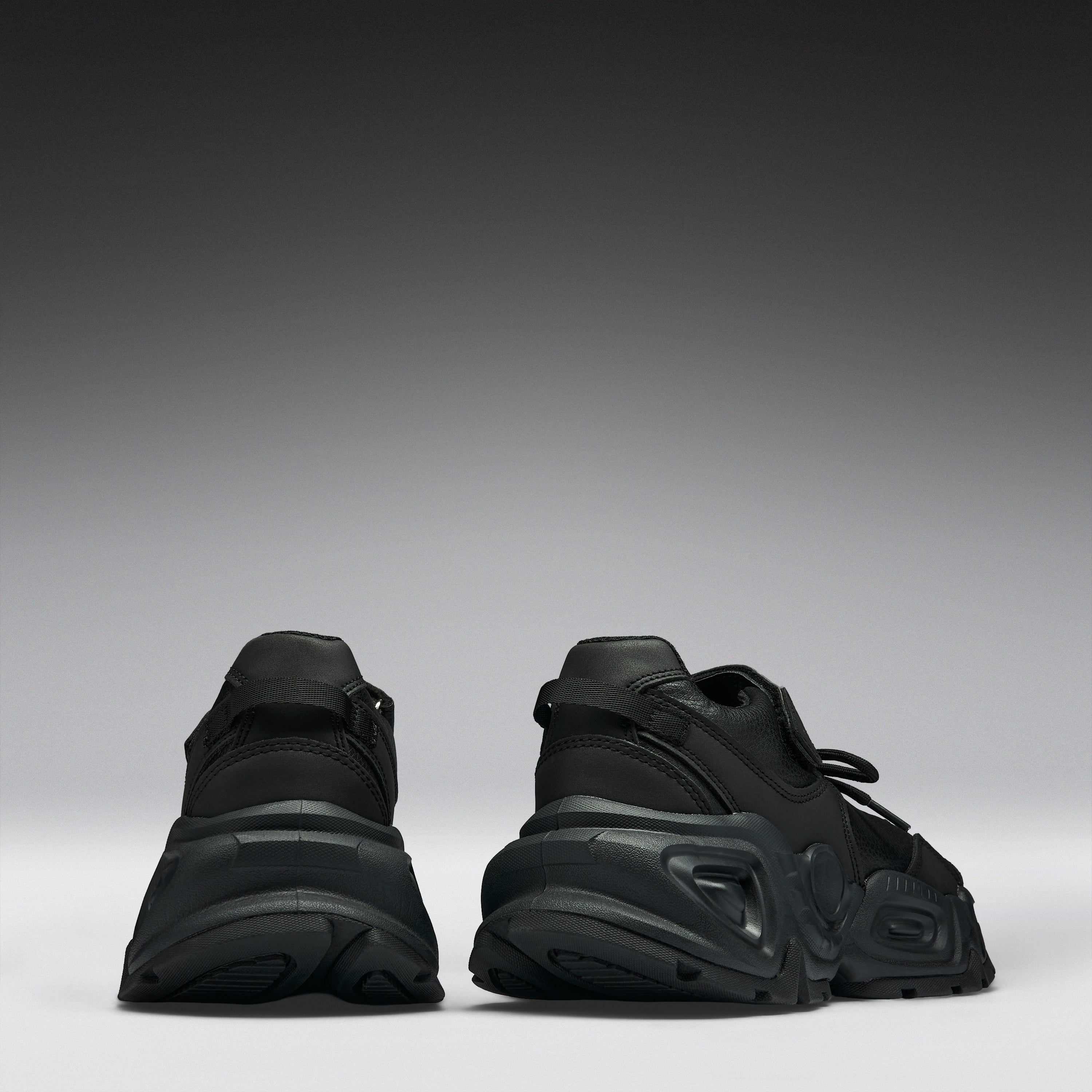 A pair of black chunky platform sneakers shown from the back, positioned side-by-side against a dark gray to light gray gradient studio background.