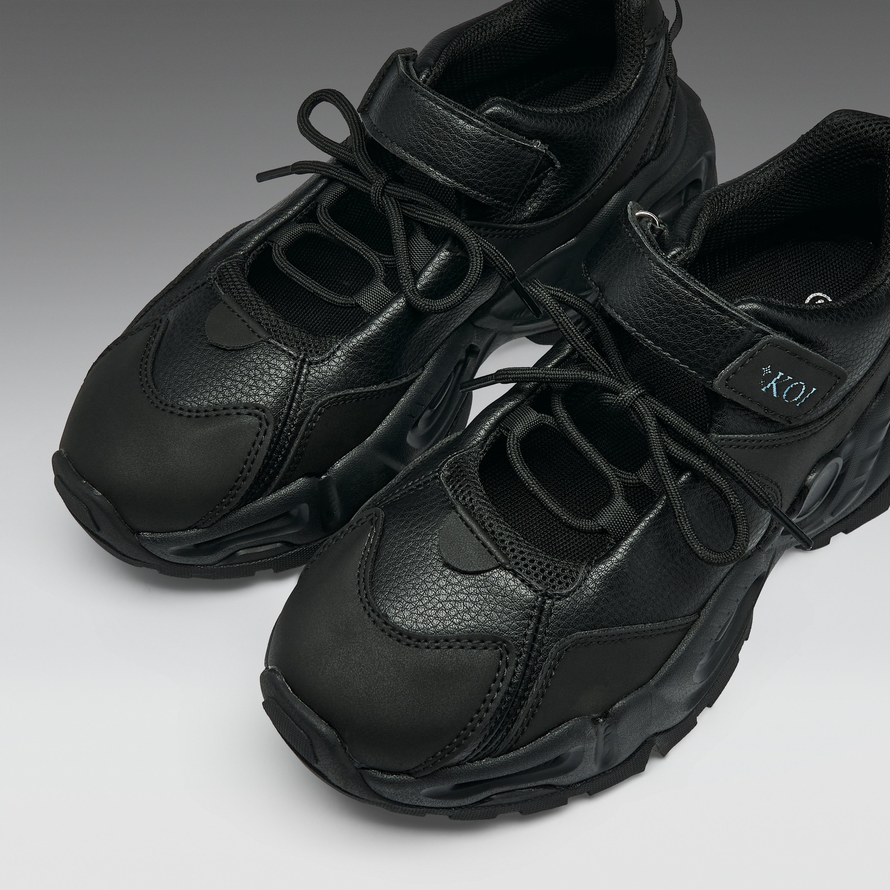 A pair of chunky black sneakers with a textured leather and mesh upper, featuring thick sculpted soles and hook-and-loop strap closures, shown on a neutral gray background.