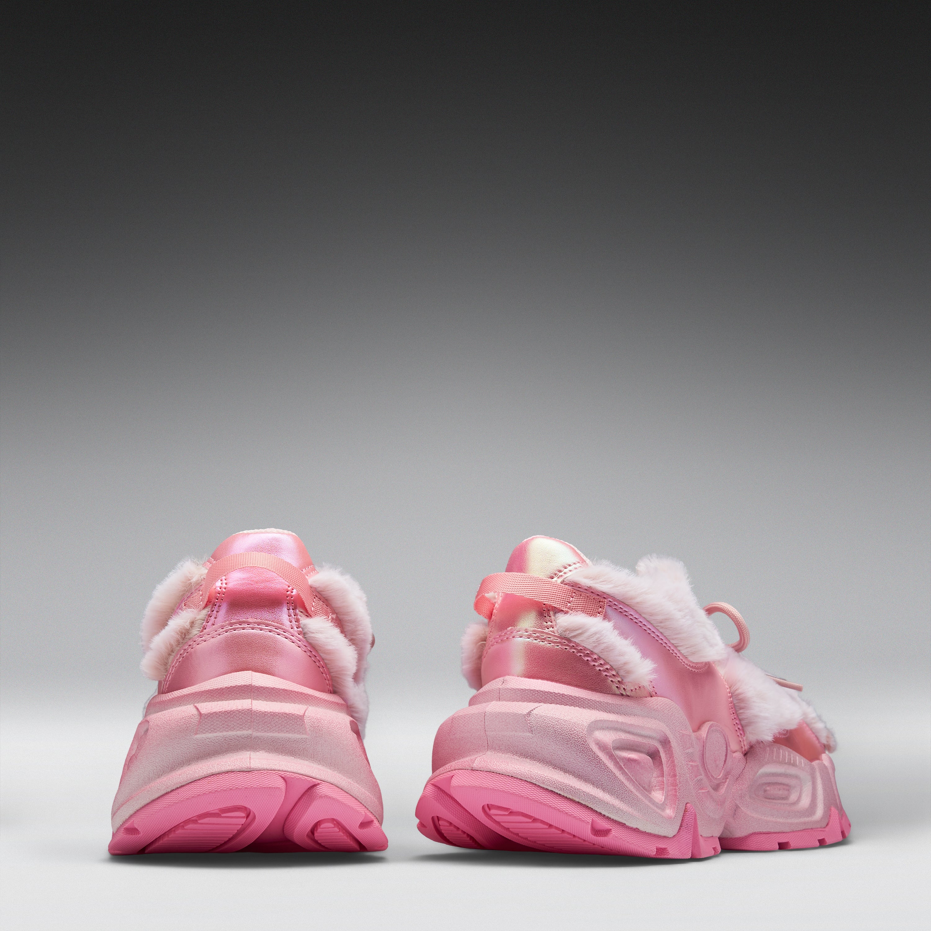 A pair of pink chunky sneakers shown from the back, featuring fuzzy white faux-fur trim and translucent pink soles, set against a dark-to-light gray gradient studio background.