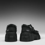 A pair of black platform sneakers shown from the back, featuring chunky lug soles, black laces, and the brand name 'KOI' printed in white on the heel.