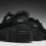 A pair of black platform sneakers with ruffled trim, studded details, and 'KOI' branding on the heel, shown against a dark gray gradient background.