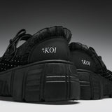 A pair of black platform sneakers with ruffled trim, studded details, and 'KOI' branding on the heel, shown against a dark gray gradient background.