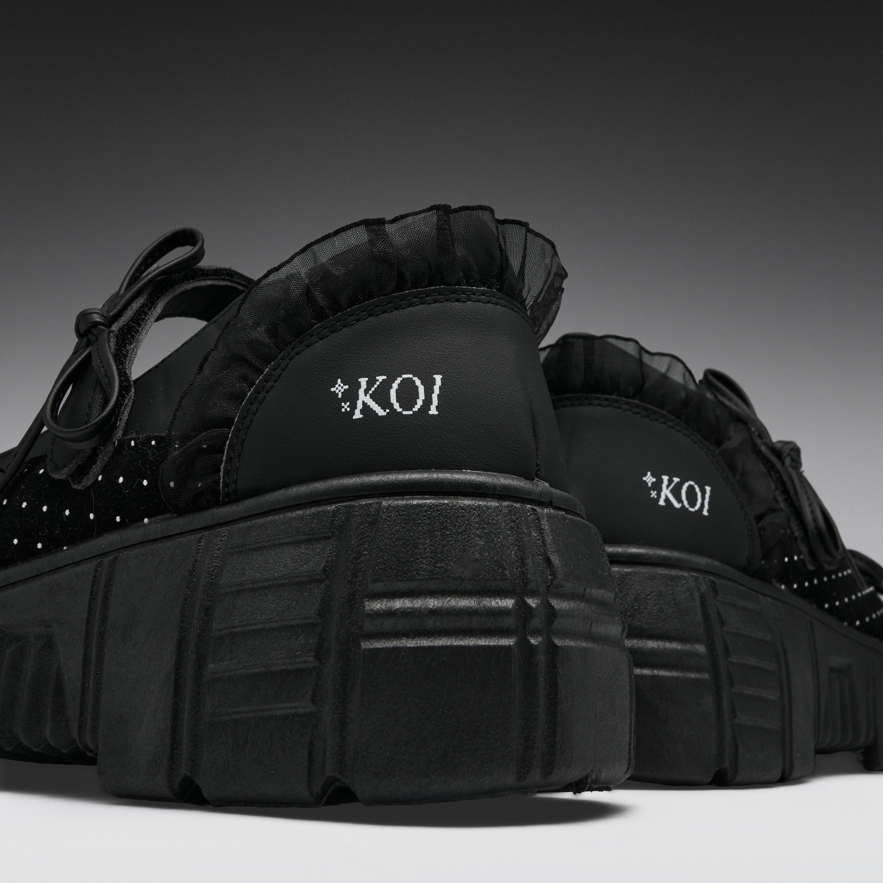 A pair of black platform sneakers with ruffled trim, studded details, and 'KOI' branding on the heel, shown against a dark gray gradient background.