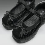 A pair of black platform Mary Jane shoes with suede toe caps, perforated details, and bow accents, shown on a neutral gray background.
