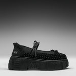 A single black platform Mary Jane shoe with a textured sole, black polka-dot toe box, and a small white brand logo on the heel, set against a dark gray gradient background.