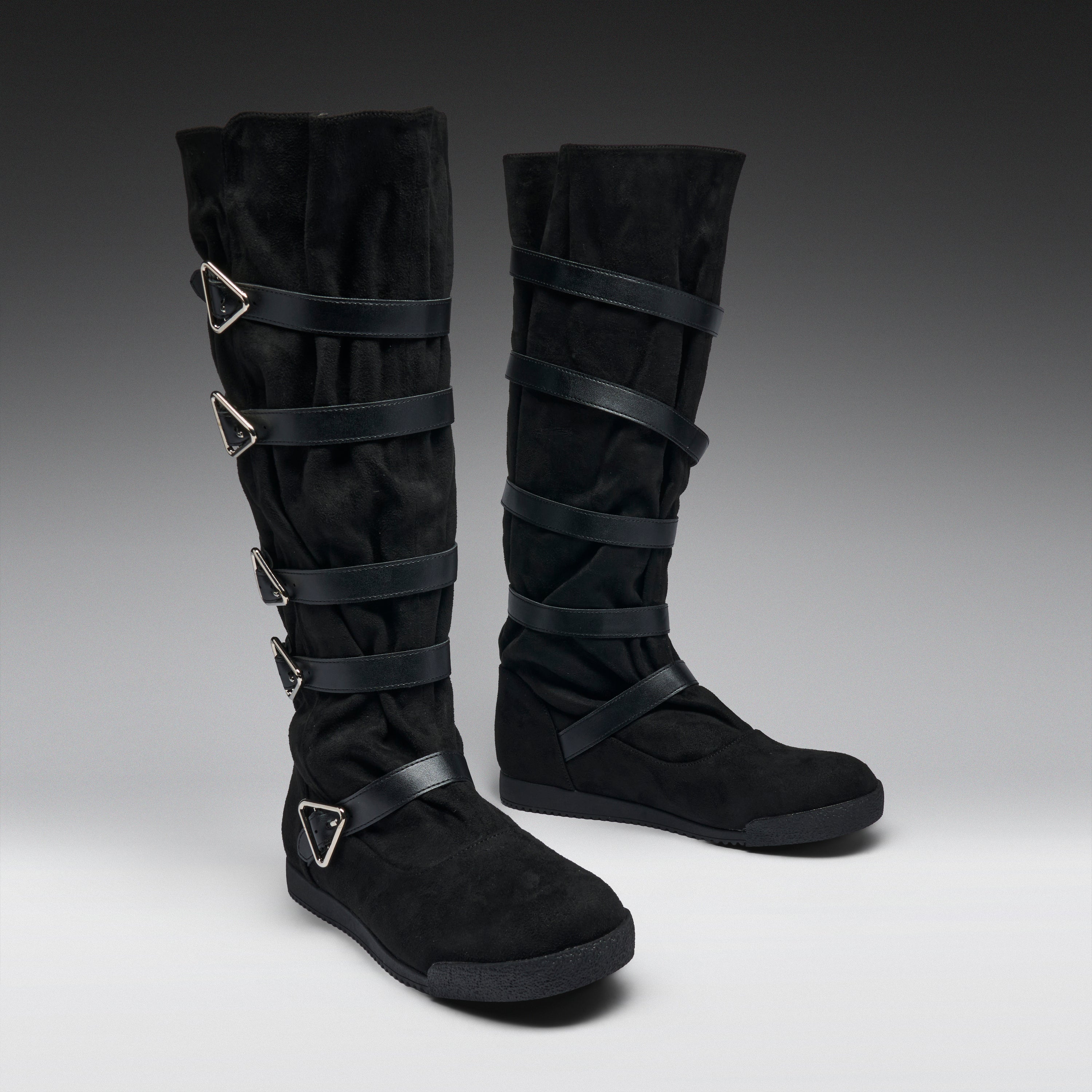 A pair of black knee-high boots with multiple wrap-around straps and silver-tone triangular buckle details, photographed on a dark-to-light gray studio background.