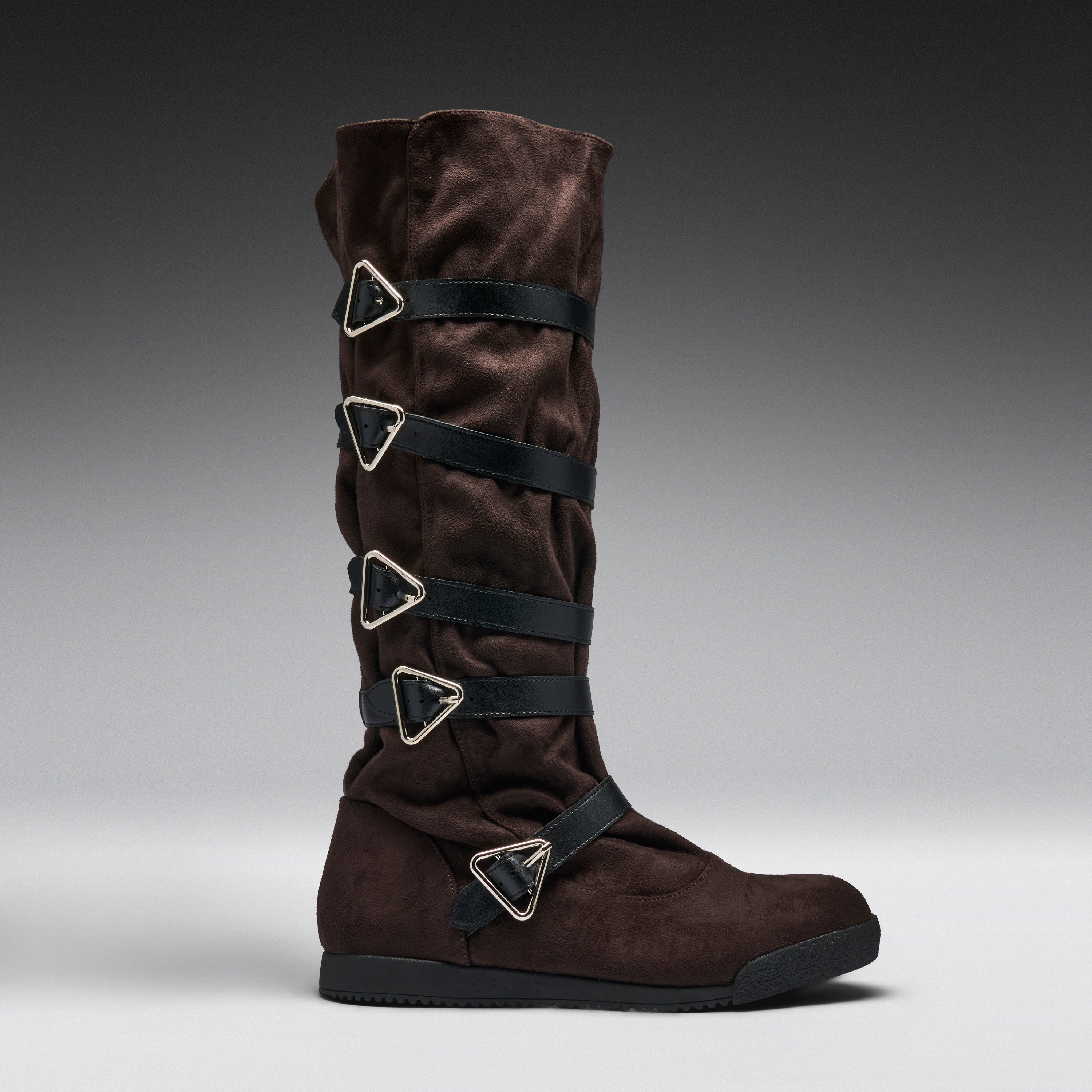 A dark brown suede knee-high boot with multiple black straps and silver-tone triangular metal hardware, shown in profile against a gray gradient studio background.