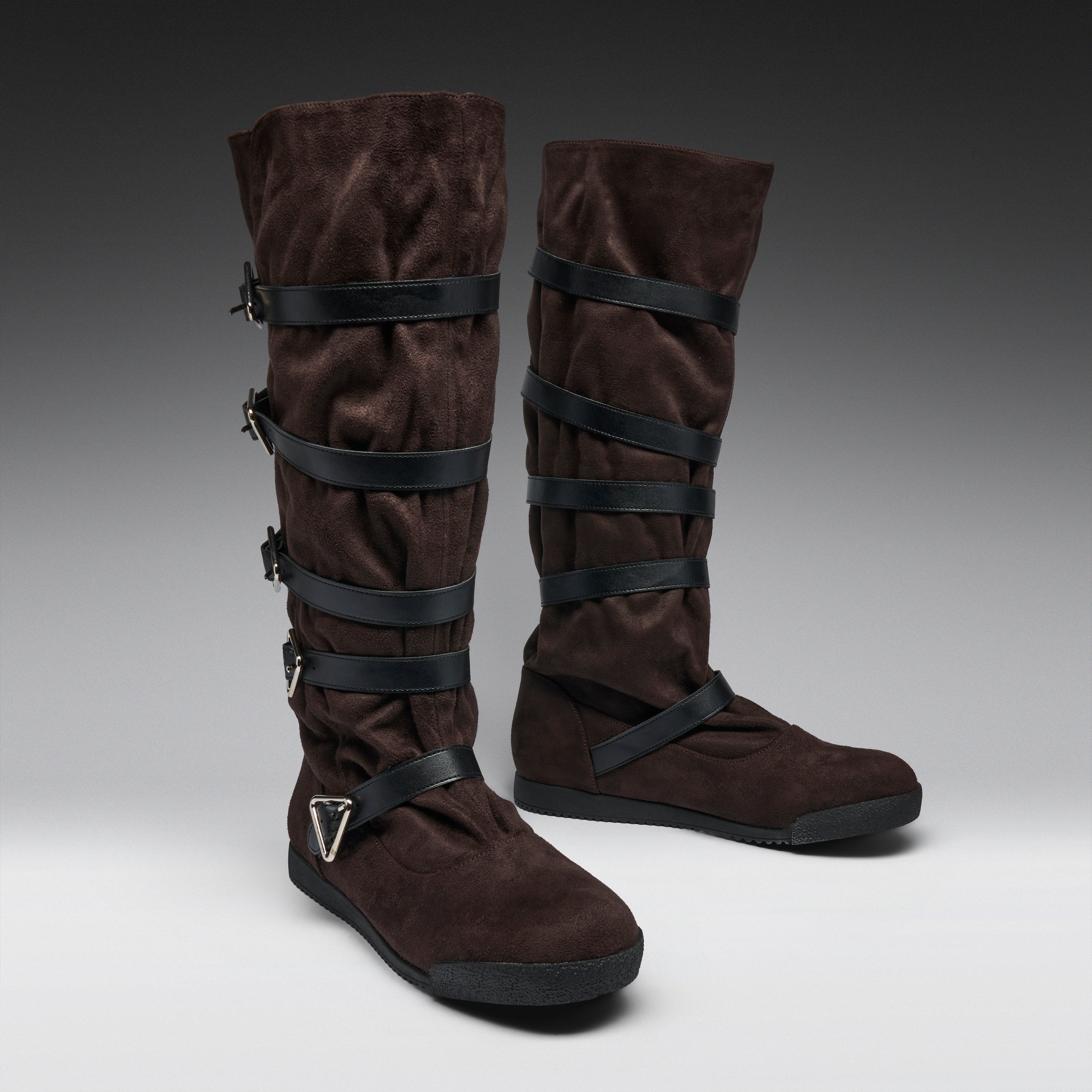 A pair of tall brown suede boots with multiple black wrap-around straps and buckles, displayed on a neutral gray studio background.