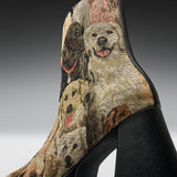 A close-up of a high-heeled ankle boot featuring a detailed woven tapestry pattern of various dogs in beige, brown, and black tones, with a solid black textured heel.
