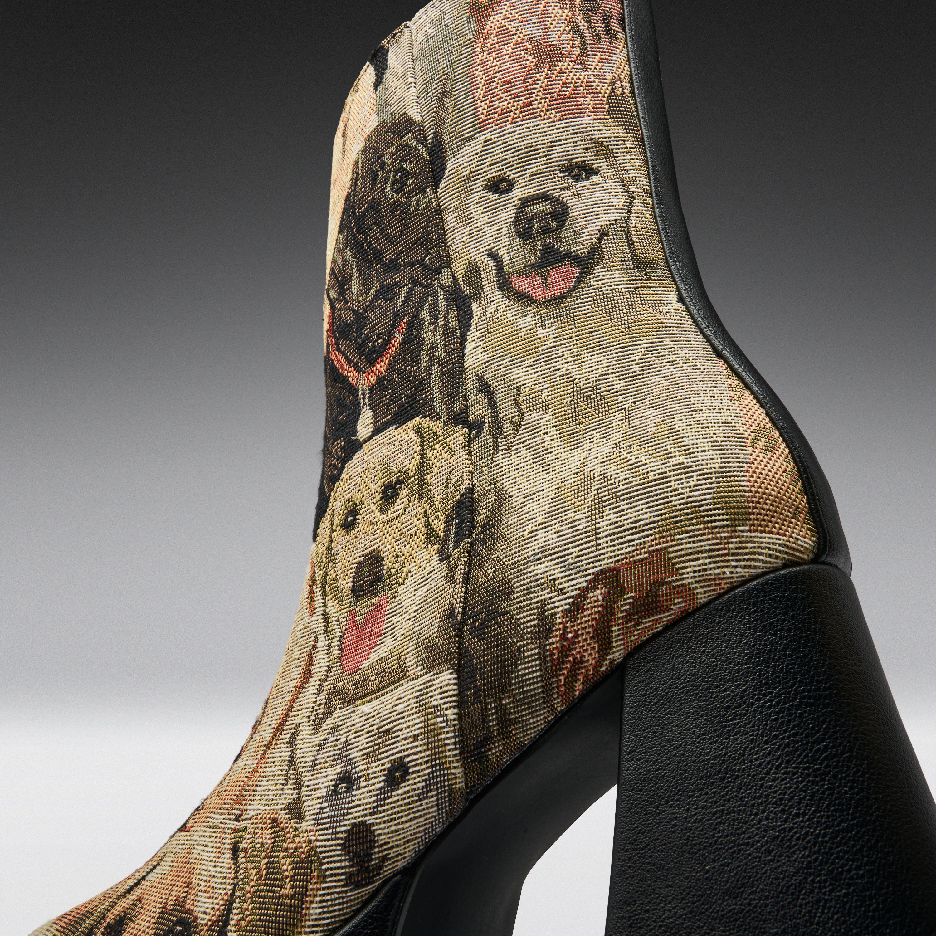 A close-up of a high-heeled ankle boot featuring a detailed woven tapestry pattern of various dogs in beige, brown, and black tones, with a solid black textured heel.