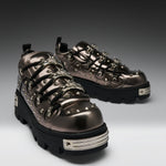 A pair of metallic bronze platform sneakers with black soles, silver studs, and decorative chain details, photographed on a dark-to-light gray gradient studio background.