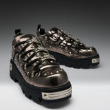 A pair of metallic bronze platform sneakers with black soles, silver studs, and decorative chain details, photographed on a dark-to-light gray gradient studio background.